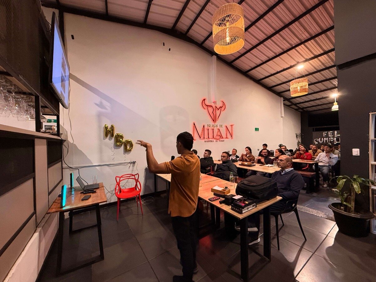 A photo capturing an indoor event or presentation where a speaker in an orange shirt addresses a seated audience in a restaurant or event space featuring a large neon sign and celebratory decorations.. Text: BRASAS DE MILAN MADRILEÑA AL FUEGO HBD FELIZ CUMPLE[AÑOS]