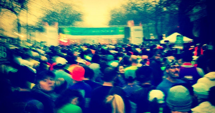 A blurry photo of a large crowd of people, likely at a race or marathon event, gathered near a banner and tents.