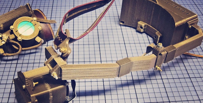 The photo shows disassembled parts of a device, possibly a VR headset or goggles, with a steampunk aesthetic. The parts appear to be 3D-printed and include a head mount, two side pieces, and a wrist-mounted component with a circular lens.