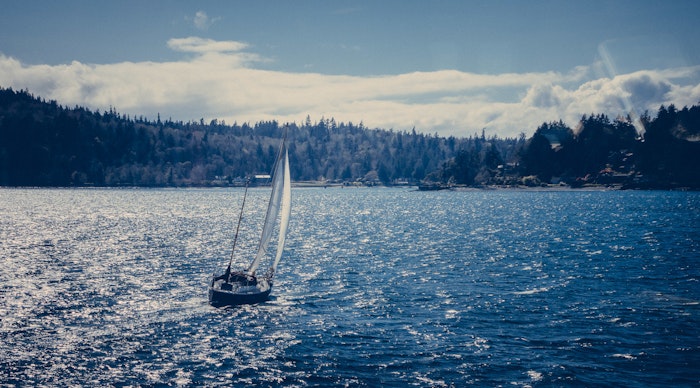 A sailboat is navigating a body of water, with a tree-lined shore in the background under a partly cloudy sky.