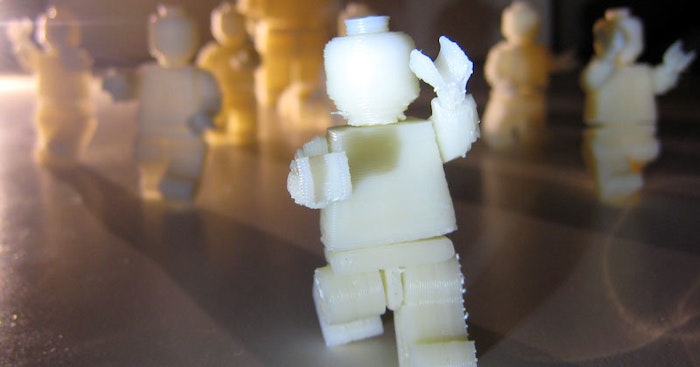 A close-up photo of a 3D-printed, white Lego minifigure, seemingly in motion, with other similar figures blurred in the background.
