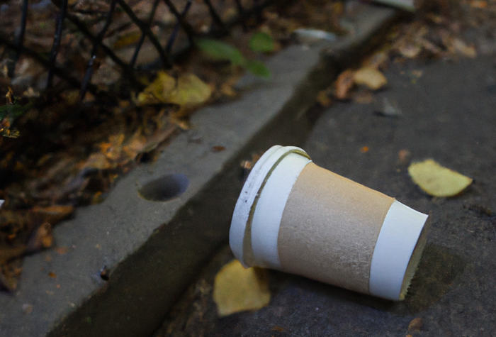A discarded paper coffee cup lies on the ground near a curb and a chain-link fence.