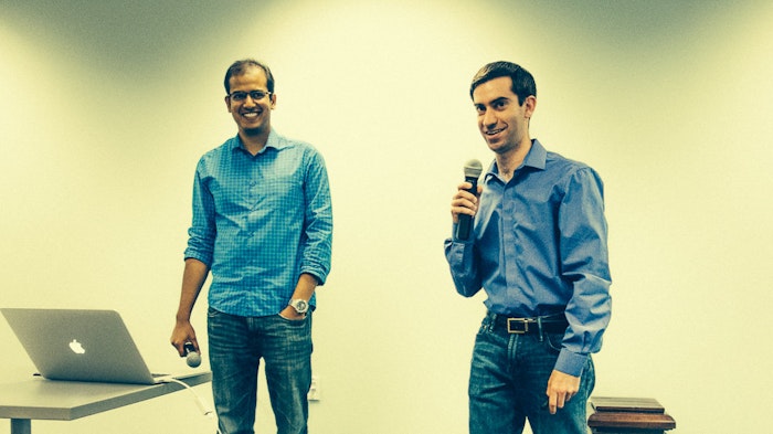 Two men stand in front of a blank wall, one holding a microphone and the other holding a microphone wire connected to a laptop on a table.