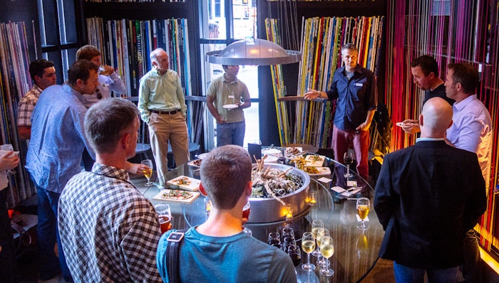 A group of people are gathered around a table with food and drinks, possibly at a social or business event. The background features a wall decorated with colorful vertical stripes.