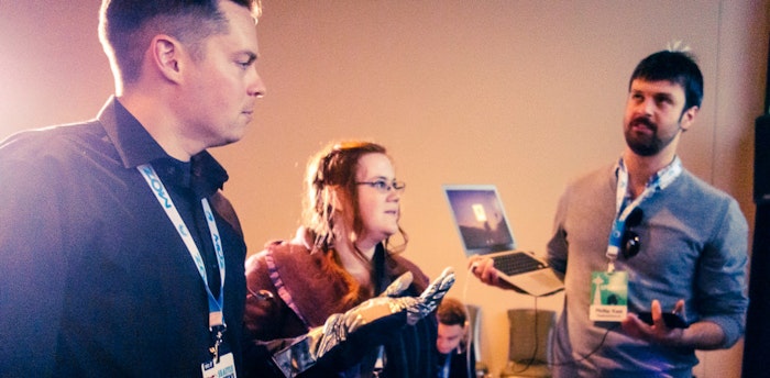 A photo of three people, two men and a woman, appearing to be at a conference or similar event. One person holds a laptop.