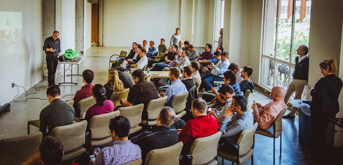 A man is giving a presentation to a group of people in a room. The audience is seated in chairs, some looking at the presenter and others looking at their phones or talking amongst themselves.
