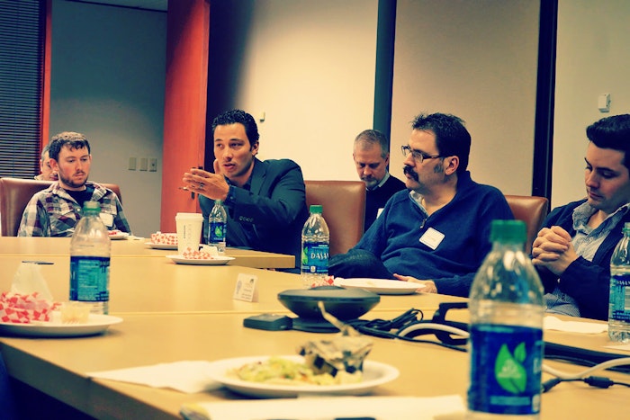 A group of people are sitting around a conference table, seemingly engaged in a discussion or meeting. One person is gesturing while speaking.