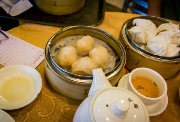 A photo depicting a table scene at a dim sum restaurant, featuring various steamed dumplings, dipping sauces, and a teapot.