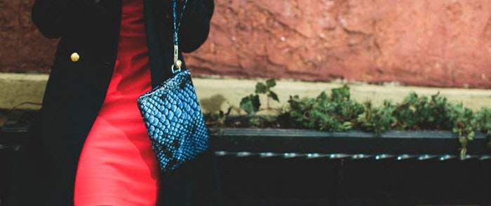 A person wearing a red dress and a dark coat is standing next to a brick wall, holding a blue textured purse.