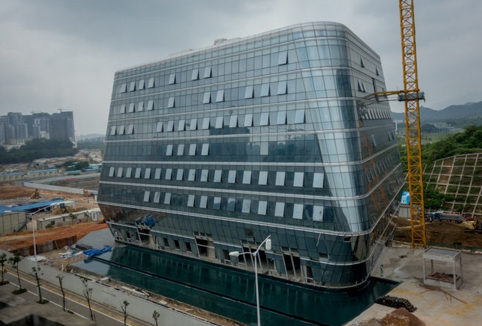 A photograph of a modern, glass-clad building under construction, with a crane visible beside it and a pool of water in the foreground.