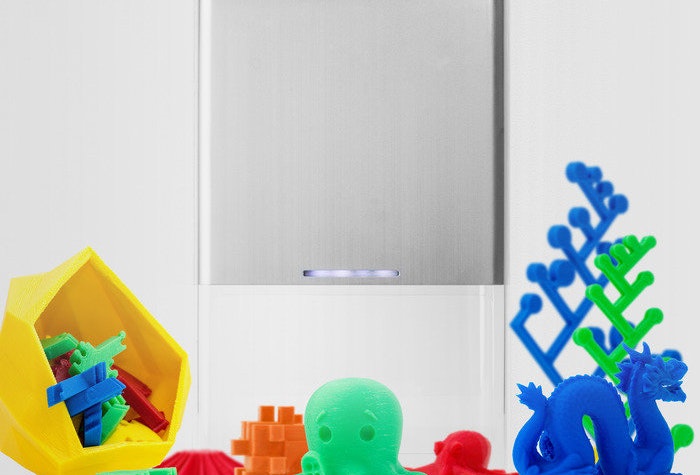 The image showcases a silver, possibly metallic, device alongside a variety of small, colorful 3D-printed objects.