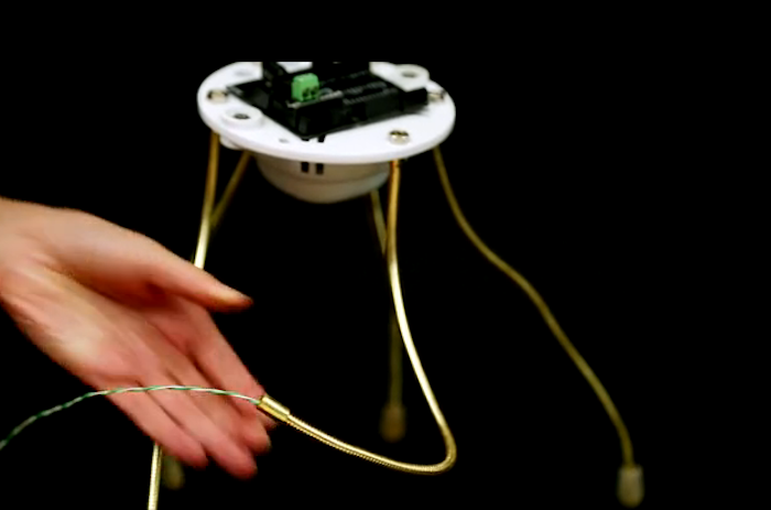 A small, white, spider-like robotic device with gold legs is shown against a black background. A hand reaches out to interact with a green and white wire connected to the device.