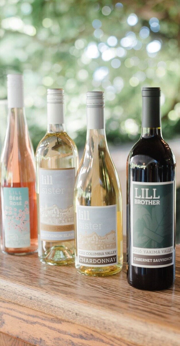 (Photo) A bright photograph featuring four different wine bottles—a rosé, two whites, and a dark red—lined up on a wooden surface against a blurred green background with bright bokeh lights. Text: LILL BÉBÉ Rosé lill sister RED MOUNTAIN SAUVIGNON BLANC lill sister 2022 COLUMBIA VALLEY CHARDONNAY LILL BROTHER 2016 YAKIMA VALLEY CABERNET SAUVIGNON Wine bottles/products | Outdoor or bright indoor setting (implied by the natural light and foliage background), on a wooden surface | Four distinct bottles with different labels (Lill Bébé, Lill Sister, Lill Brother), shallow depth of field (bokeh background) | Product photography/lifestyle shot | Colors: #A5927B, #4F6D55, #F0EFEA, #402830 Note: The image is a realistic capture of physical objects (wine bottles) in a specific setting, characteristic of commercial or lifestyle product photography.