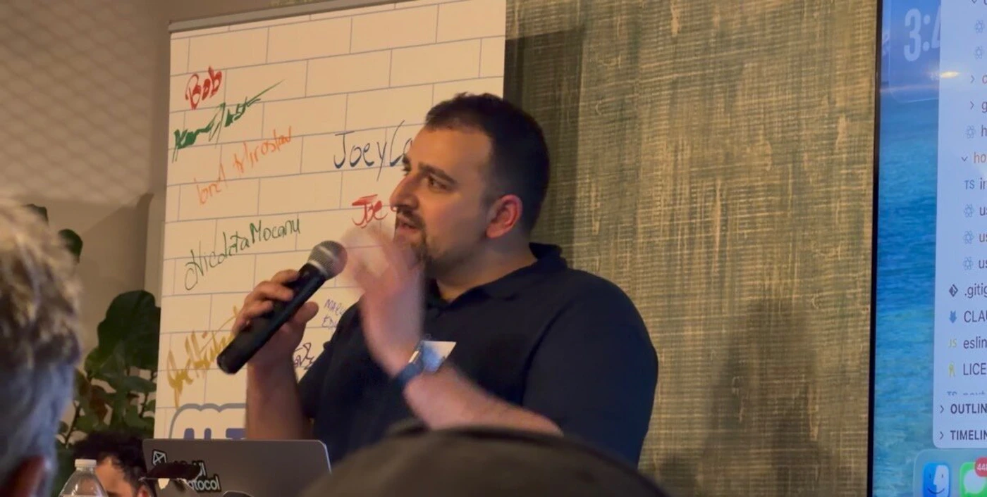 (Photo) A man is giving a presentation or talk indoors, holding a microphone and gesturing, standing in front of a whiteboard covered in names and next to a screen displaying programming code. Text: Bob, [unreadable signature], brd [unreadable], Nicolae Mocanu, Joey Co[unreadable], Je[unreadable], 3:4[x], .gitig[x], CLAU[x], eslint[x], LICEN[x], OUTLINE, TIMELINE. Presentation/speaker event | indoor (meetup or conference room) | Speaker holding mic, whiteboard with writing, visible code screen, audience members in foreground. | Candid, event photography | Colors: #292931, #FFFFFF, #808080, #50A0B0 Note: The image is a photograph capturing a real-world, candid moment of a speaker at an indoor technical or community event.