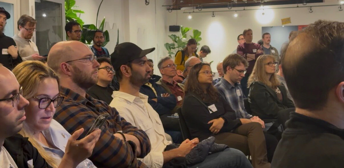 (Photo) A candid photograph captures a diverse audience of approximately twenty people seated and standing indoors, all focused intently on an unseen speaker or presentation area. Group of people attending an event/meetup | indoor (event space or office) | Large group, many attendees wearing name tags, audience is focused forward | candid, event photography | Colors: #4F382E, #FFFFFF, #222222, #A0A0A0 Note: The image is a realistic capture of a real-world event featuring a large group of people in an indoor setting, fitting the definition of event photography.