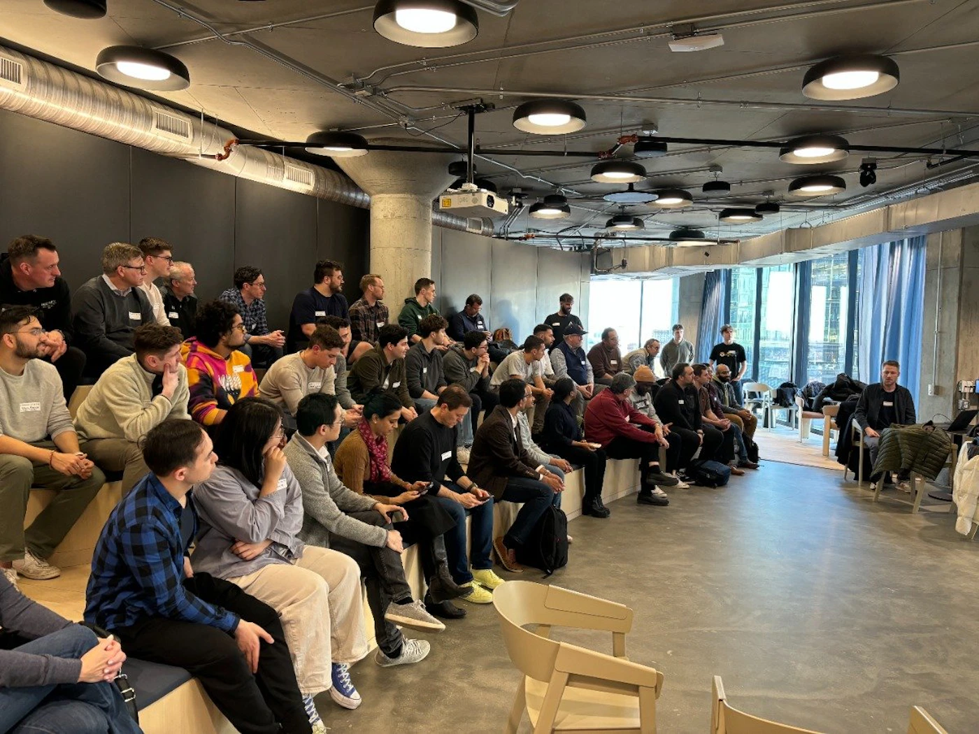 (Photo) A photo of a group of people sitting in tiered seating in a large room, appearing to be listening to a speaker. people | indoor | group of people, presentation setting | candid Note: The image is a real-life captured image of a group of people in a specific setting, indicating it is a photograph.