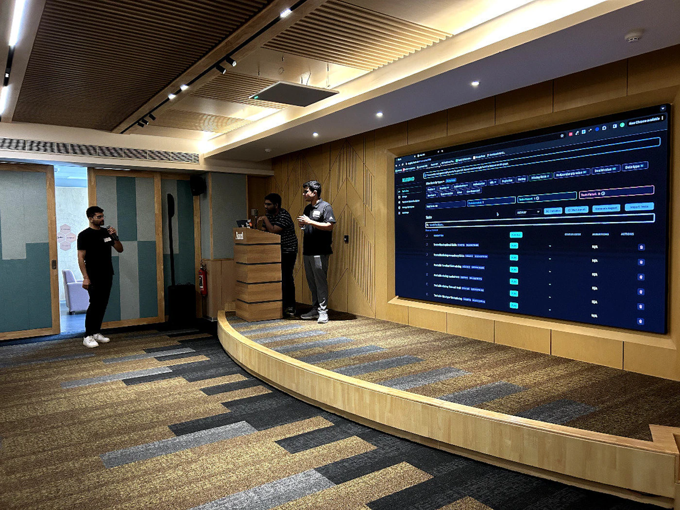 (Photo) A photo of a presentation taking place in a modern conference room. Three individuals are on a small stage, one speaking into a microphone, while a large screen displays a spreadsheet or table of data. people, presentation | indoor | presentation screen, stage, audience | candid Note: The image is a real-world captured image of a scene with people and a screen, making it a photograph. It's not a logo, illustration, banner, or handwriting.