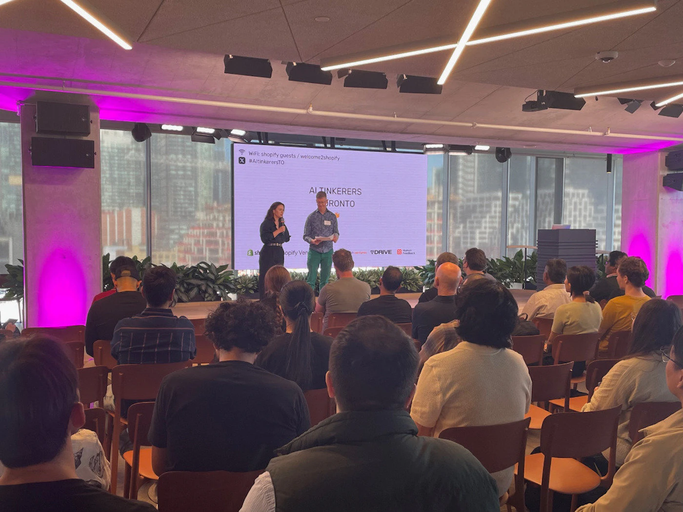 (Photo) A photo of a presentation taking place in a modern office space with a large audience. Text: We're shopify partners / welcome2shopify #AltTinkersTO ALTINKERS TORONTO Apply Venue TORVE people | indoor | presentation, audience, modern office | candid Note: The image is a real-life capture of an event, depicting a presentation with an audience in a physical space. This makes it a photograph.