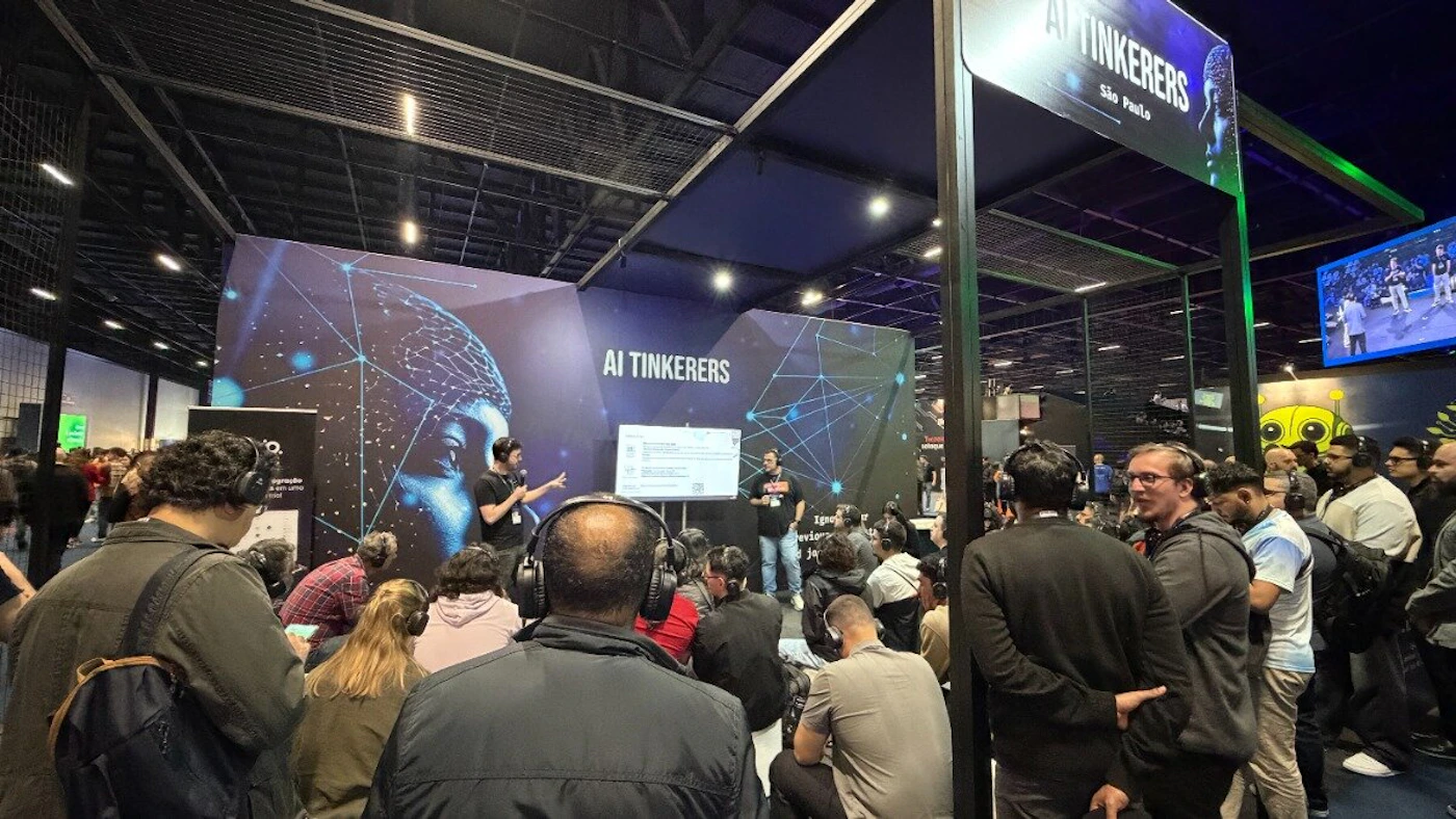 (Photo) A photograph captures an indoor event, likely a trade show or convention, where two presenters are conducting a workshop in front of a large screen branded 'AI TINKERERS'. A crowd of attendees is seated on the floor, many wearing headphones, suggesting a silent presentation or audio-assisted instruction. Text: AI TINKERERS São Paulo Q. Event attendees, presenters, and a branded workshop booth/stage focused on AI. | Indoor convention center or large event hall, characterized by high ceilings and dark lighting. | Audience members wearing headphones, large digital backdrop featuring abstract network/brain graphics, and industrial ceiling structure. | Candid, environmental shot. | Colors: #1A1A1A, #000033, #FFFFFF, #33A0FF Note: The image is a photograph capturing a real-world scene, specifically an environmental shot of an event taking place inside a large hall with people, staging, and branding.