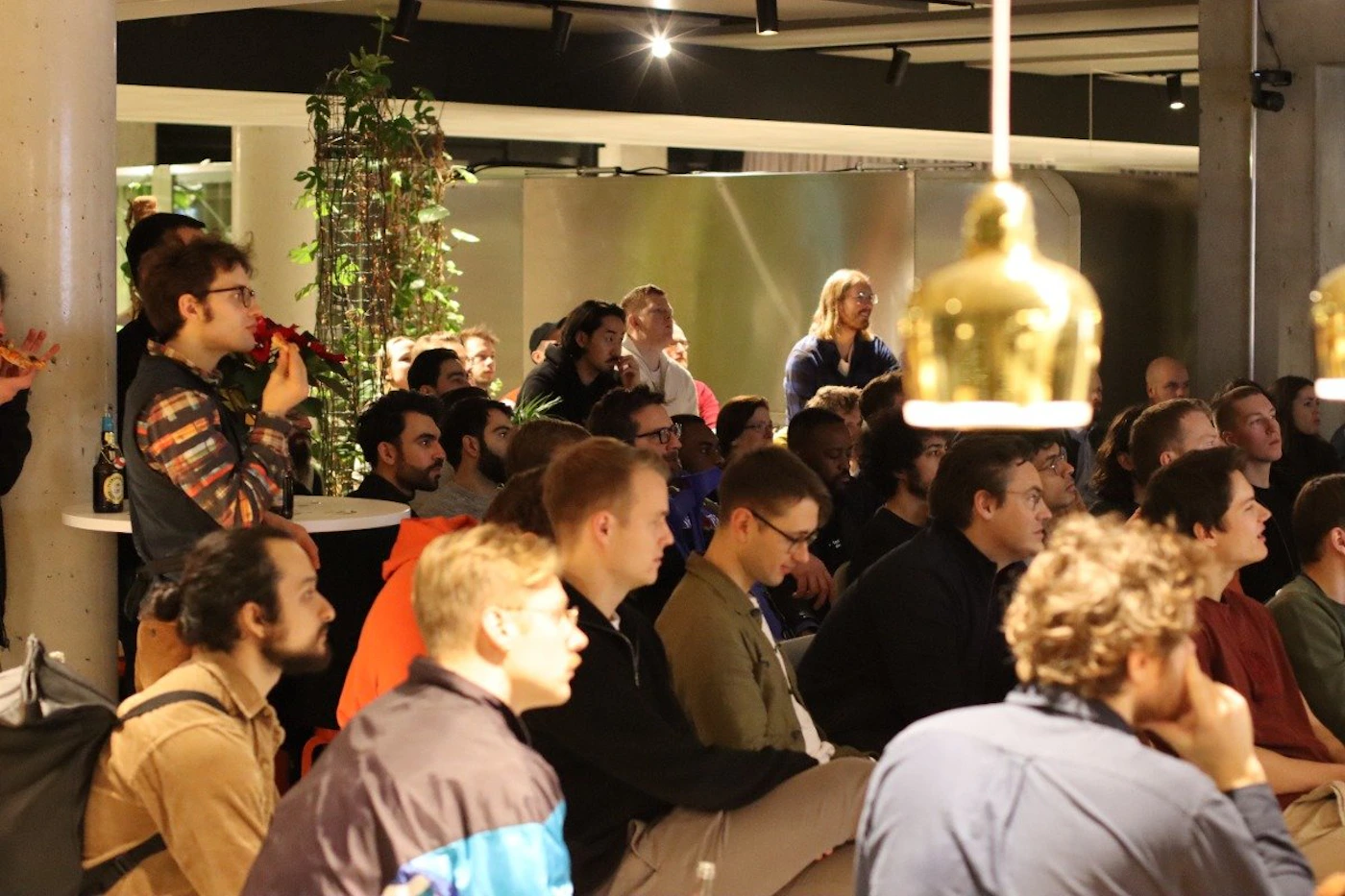 (Photo) A photo of a group of people sitting and standing in an indoor setting, appearing to watch a presentation or performance. people | indoor | group of people, casual setting, presentation/performance implied | candid Note: The image is a real-life captured moment of a group of people in a casual indoor environment, indicative of a photographic snapshot.