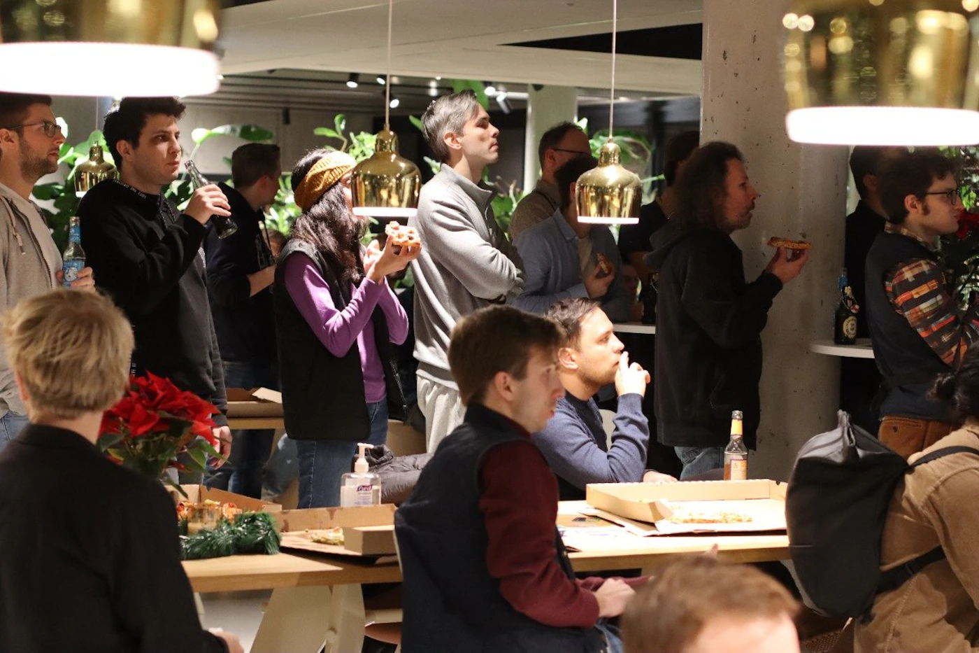 (Photo) A group of people are gathered in a brightly lit space, eating pizza and socializing.  They are standing and sitting around tables. people | indoor | group of people, food, social gathering | candid Note: The image is a candid photograph of a real-life scene, capturing a moment in time.