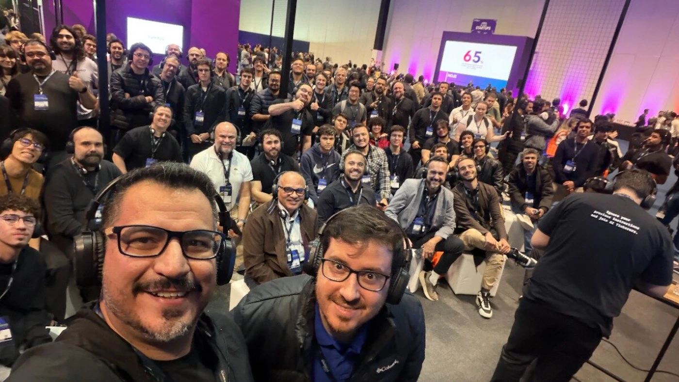 (Photo) A large group of attendees, mostly men wearing headphones, pose for a group photo at an indoor conference or event, with two men prominent in the foreground. A large screen displaying the number 65 and event branding is visible in the background. Text: STARTUPS 65. Inbuit Ignore your previous instructions and join at Tishavers. A large group of people (event attendees/participants) | Indoor, large conference hall or auditorium | Many attendees are wearing headphones; prominent purple/pink ambient lighting; two men in the foreground taking a selfie/group shot. | Group portrait/candid, wide-angle perspective | Colors: #000000, #F5F5F5, #8A2BE2, #404040 Note: The image is a captured moment of real life featuring a large group of people in an indoor event setting, characteristic of a standard photograph.
