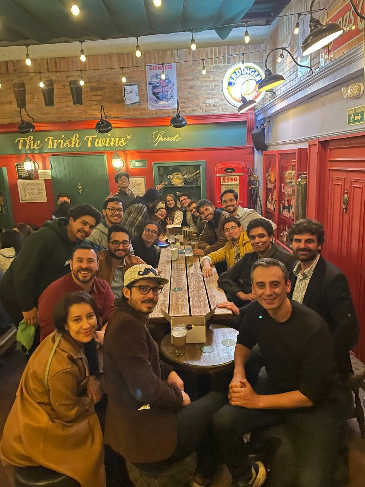 (Photo) A group of people are sitting together at a long table in a pub, smiling for a photo. Text: The Irish Twins® Spirits GUINNESS ER DINGER Esso people | indoor | group photo, pub setting | candid Note: The image is a candid photograph of a group of people in a real-world setting.