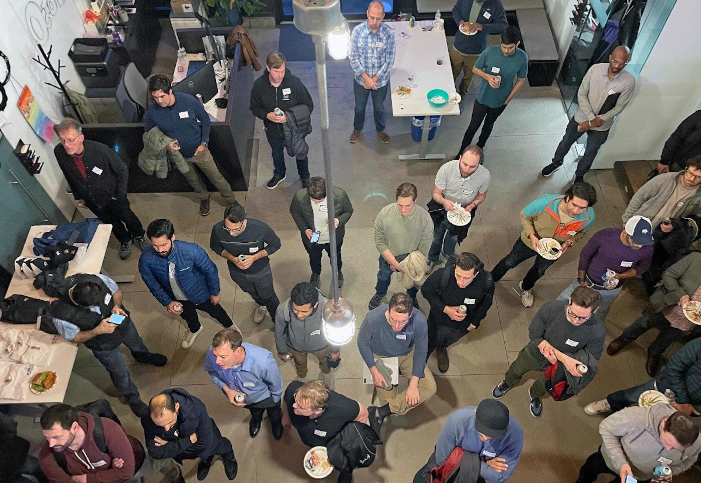 (Photo) A high-angle photo of a group of people gathered in an office space, many holding drinks and plates of food. people | indoor | group of people, office setting, food and drinks | candid Note: The image is a real-life captured moment of people in an office setting, making it a photograph. It's not a drawing, logo, or other created graphic.