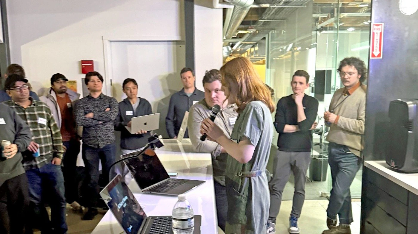 (Photo) A woman speaks into a microphone while giving a presentation to a small group of people in an office setting.  Two laptops are visible on a table in front of her. people | indoor | presentation, group of people, laptops | candid Note: This is a real-life captured image of a scene with people, depicting a specific event.  It's not a drawing, logo, or banner.