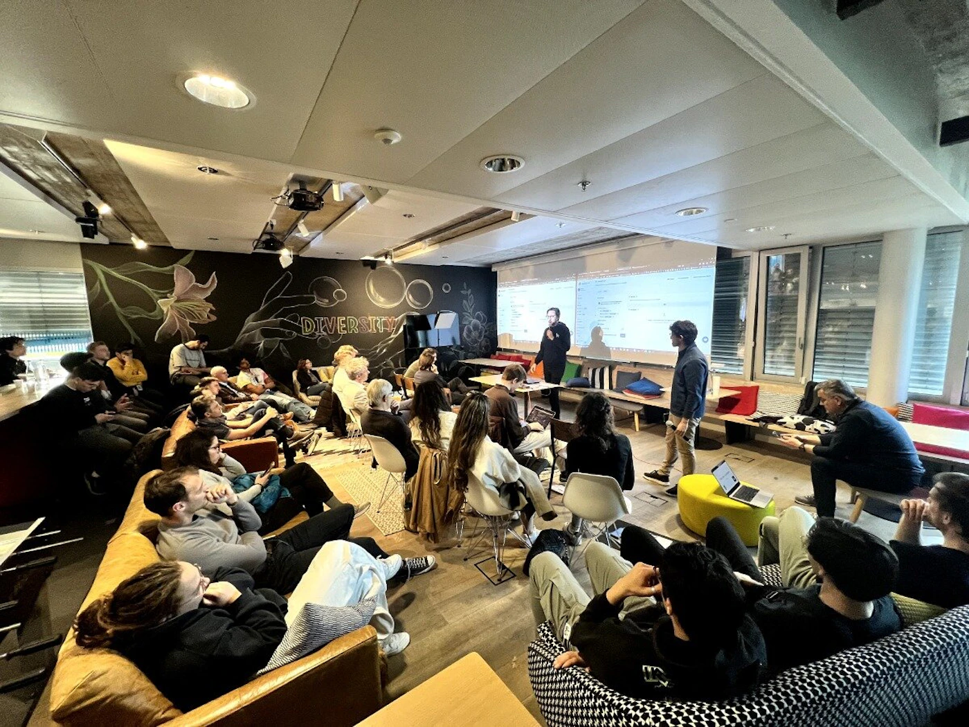 (Photo) An indoor photograph captures a diverse group of people gathered in a modern, casual setting, likely attending a presentation or workshop. A large screen displays content, and a speaker stands near it, while attendees are seated on sofas, beanbags, and chairs. Text: DIVERSITY [unreadable text on screen] group of people, presentation/event | indoor, modern lounge/meeting space | projector screen, wall art, casual seating (sofas, beanbags), multiple attendees, speaker | candid, event photography | Colors: #8B4513, #000000, #FFFFFF, #F5F5DC Note: This image is a direct capture of a real-world scene, featuring people and an environment, characteristic of a photograph.