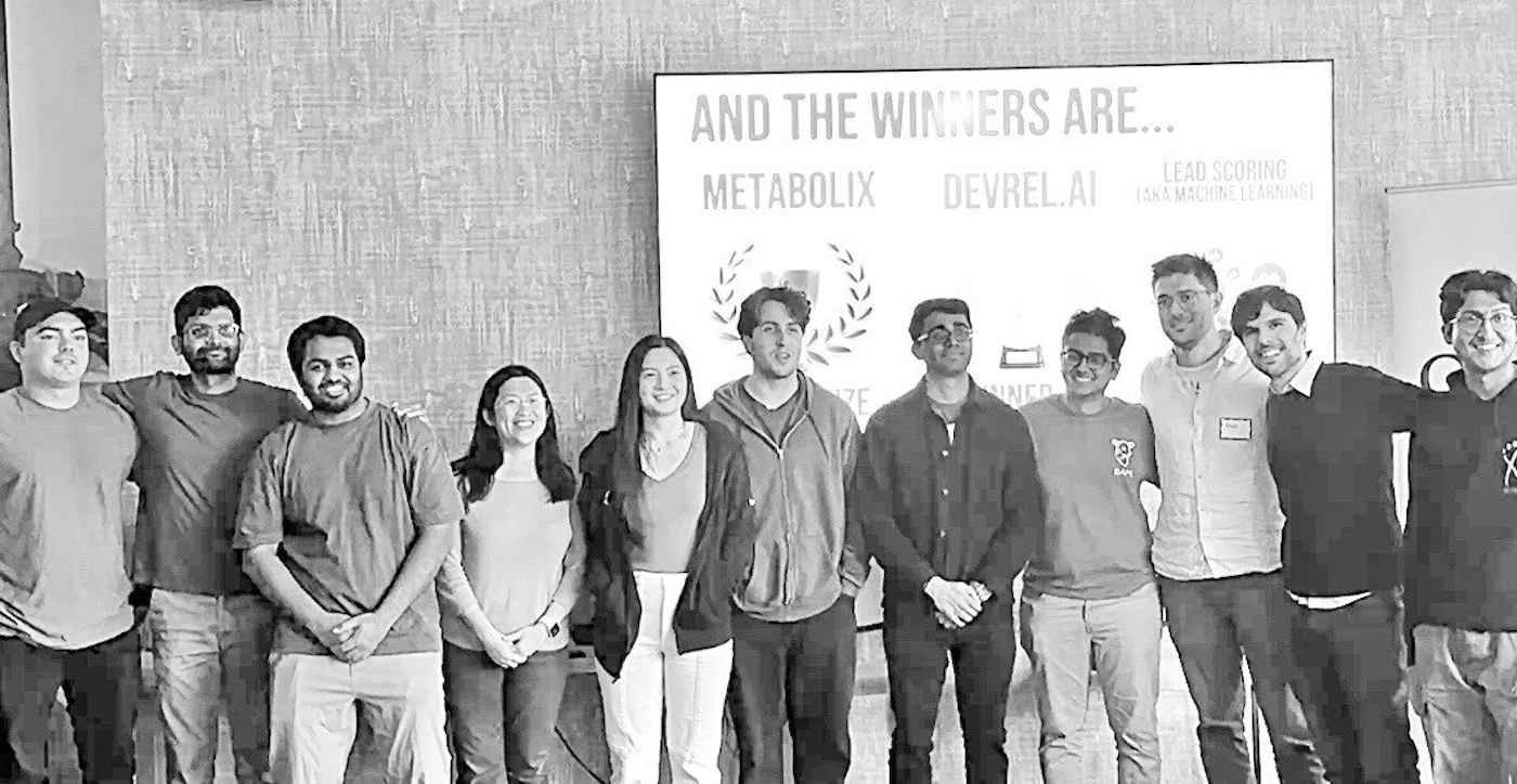 (Photo) A black and white photo of a group of people standing together, possibly award winners based on the text on the screen behind them. Text: AND THE WINNERS ARE... METABOLIX DEVREL.AI LEAD SCORING (VIA MACHINE LEARNING) people | indoor | group photo, award winners | candid Note: The image is a real-life captured image of a group of people, making it a photograph. The presence of a screen with text suggests an award or announcement scenario.