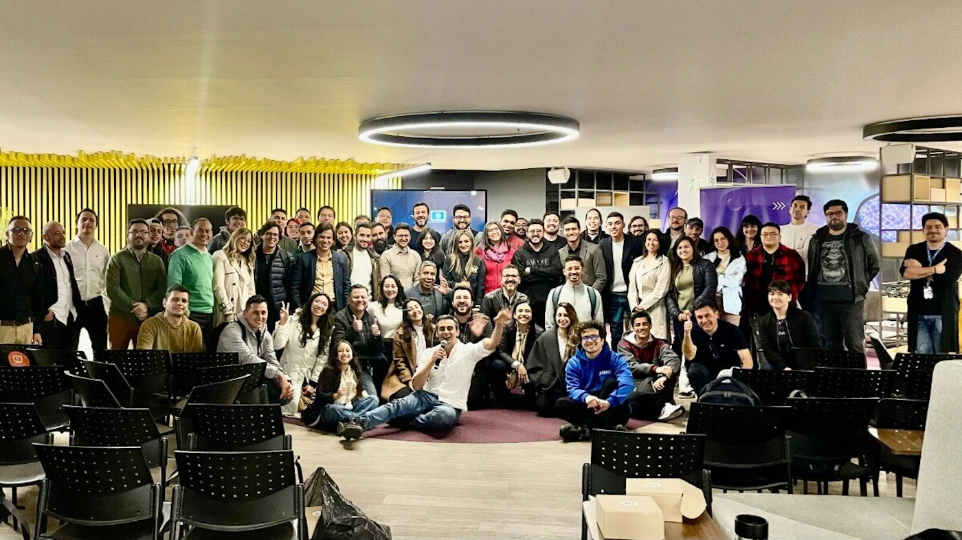 (Photo) A large group of people is posing for a photo in a modern office or event space. Some are standing, some are sitting on the floor, and empty chairs are visible in the foreground. people | indoor | large group, office setting | group photo Note: This is a photograph capturing a group of people in a specific setting. It's not a logo, illustration, banner, or handwriting.