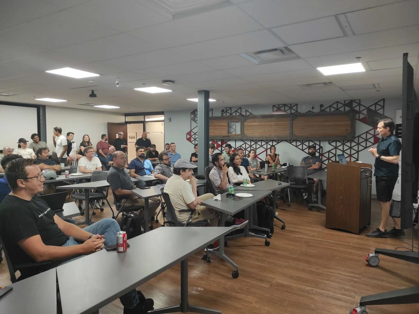 (Photo) A man is presenting to a group of people seated at tables in a room. people | indoor | presentation, audience, tables, chairs | candid Note: The image is a real-life captured scene of a presentation, depicting people and a room, making it a photograph.