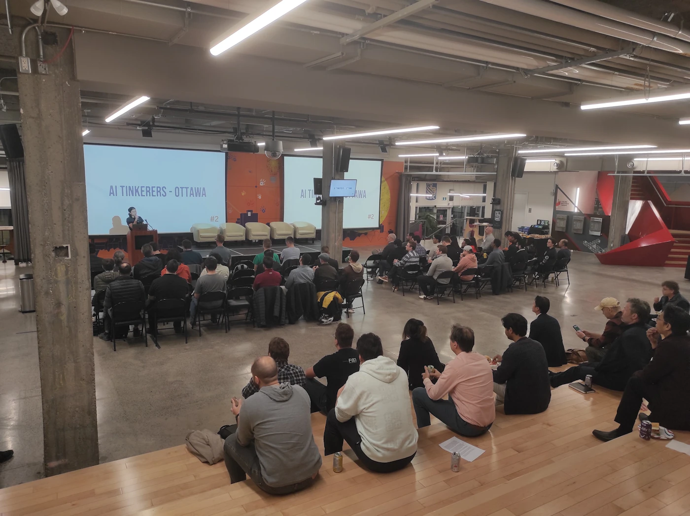 (Photo) A photo of a presentation or meeting taking place in a large, modern space.  An audience sits facing a speaker and two large screens. Text: AI Tinkeres - Ottawa #2 AI Tinkering - Ottawa people | indoor | presentation, audience, large screens | candid Note: The image is a real-life captured image of an event, depicting people in a presentation setting. This clearly falls under the category of a photograph.