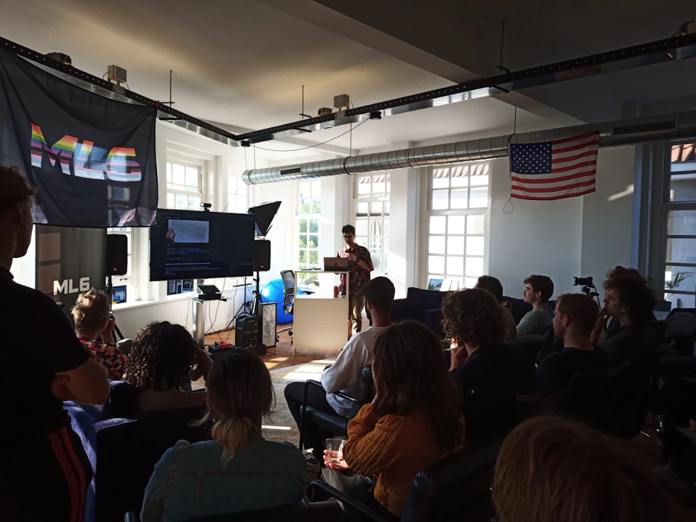 (Photo) A person is giving a presentation to a small audience in a room.  A screen displays information, and flags hang from the ceiling. Text: ML6 people | indoor | presentation, audience, flags | candid Note: The image is a real-life captured scene of an event, depicting people and their environment. This makes it a photograph.