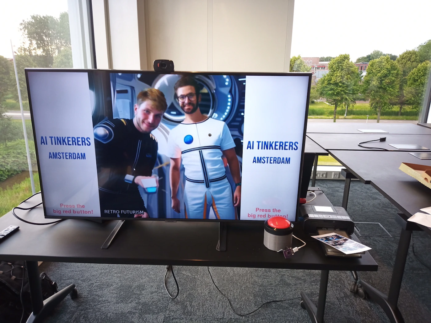 (Photo) A photo of a television screen displaying two men in futuristic attire, with text on the screen and a large red button in front of the TV. Text: AI TINKERERS AMSTERDAM Press the big red button! RETRO FUTURISM AI TINKERERS AMSTERDAM Press the big red button! screen display, people, indoor space | indoor | television screen, futuristic costumes, red button | candid Note: The image is a real-life photograph of a screen, a button, and a room. It captures a scene rather than representing a logo, banner, or other graphic element.