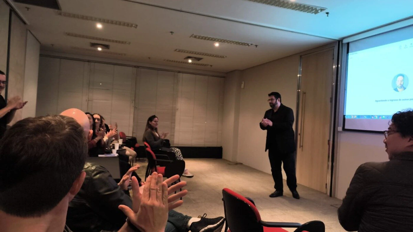 (Photo) The image captures an indoor scene of a conference or presentation, where a man in dark clothing is standing at the front, addressing an audience. Several people in the audience are seen clapping, and a projector screen displays some text and a profile icon. Text: Aguardando o ingresso de outros p People, presentation, meeting | Indoor, conference room/lecture hall | A speaker/presenter, an audience clapping, a projector screen with text and an icon. | Candid, event photography | Colors: #363636, #D3CFCB, #B22222, #ADD8E6 Note: This image is a direct capture of a real-world event or scene, featuring people and an environment, which aligns with the definition of a photograph rather than an illustration, logo, or banner.
