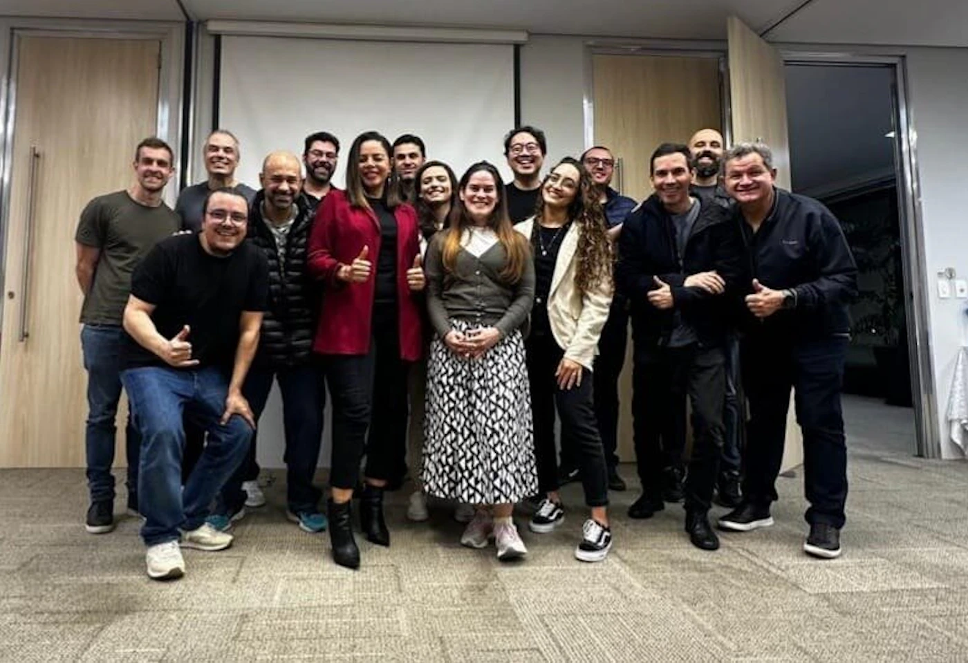 (Photo) The image features a diverse group of approximately 15 people, both men and women, posing together in what appears to be an indoor office or conference room setting. Most individuals are smiling and some are giving a thumbs-up gesture, suggesting a positive and collaborative atmosphere. group of people | indoor, office/conference room | multiple people, smiling, some giving thumbs up, diverse group, casual to business-casual attire | group portrait, candid | Colors: #2A2A2A, #7A7A7A, #D4C7B5, #A32A3B, #D9D2C6 Note: The image is a direct capture of real people in a real-world setting, characteristic of a photograph. It is not a drawing, logo, banner, or handwriting.