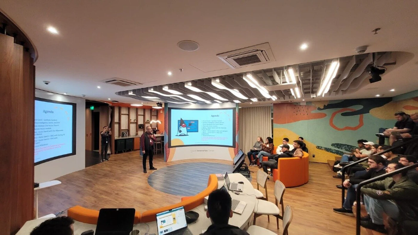 (Photo) A wide-angle photograph captures an indoor presentation or lecture in a modern, well-lit room. A speaker stands at the front, facing an audience seated in various arrangements, while two large screens display presentation slides. Text: Agenda Agenda Oracle Innovation Lab People attending a presentation or lecture | Indoor, modern conference room or auditorium | A speaker at the front, an audience seated in tiered rows and lounge chairs, two large projection screens displaying 'Agenda' slides, distinctive wavy ceiling lights, and colorful abstract wall art. | Candid, wide-angle shot of an event | Colors: #FF8C00, #8B4513, #4682B4, #F5F5DC Note: This image is a direct capture of a real-world scene, showing people, an indoor setting, and an event in progress, which are characteristics of a photograph.