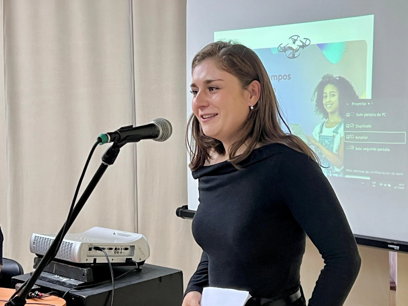 (Photo) A woman with long brown hair, wearing a black top, smiles while speaking into a microphone in an indoor setting. Behind her, a projector displays a presentation slide featuring text, options for screen projection, and an image of a young girl. Text: mpos Proyectar Solo pantalla de PC Duplicado Ampliar Solo segunda pantalla Más opciones de configuración A person (woman) giving a presentation or speech. | Indoor, likely a conference room or classroom. | A microphone on a stand, a white projector, and a large screen displaying a presentation with text and an image of a young girl. | Candid, event photography. | Colors: #1A1A1A, #E8E5DF, #B0C0E0, #E0E0E0 Note: The image is a realistic depiction of a scene captured by a camera, showing a person and objects in a specific environment, characteristic of a photograph.