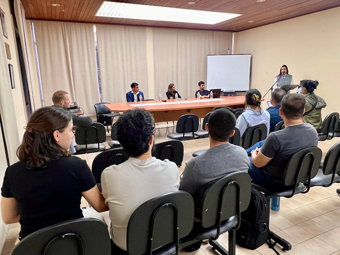 (Photo) The image shows an indoor meeting or lecture taking place, with an audience seated in rows facing a speaker at a podium and a panel seated at a long table. A white projection screen is visible behind the speaker. people attending a presentation or meeting | indoor, conference room or lecture hall | multiple people, a speaker at a podium, a panel table, a projection screen, wooden ceiling | candid, documentary | Colors: #e0d9c8, #8b5a2b, #333333, #ffffff Note: This image is a realistic capture of a real-world scene, characteristic of a photograph taken with a camera.