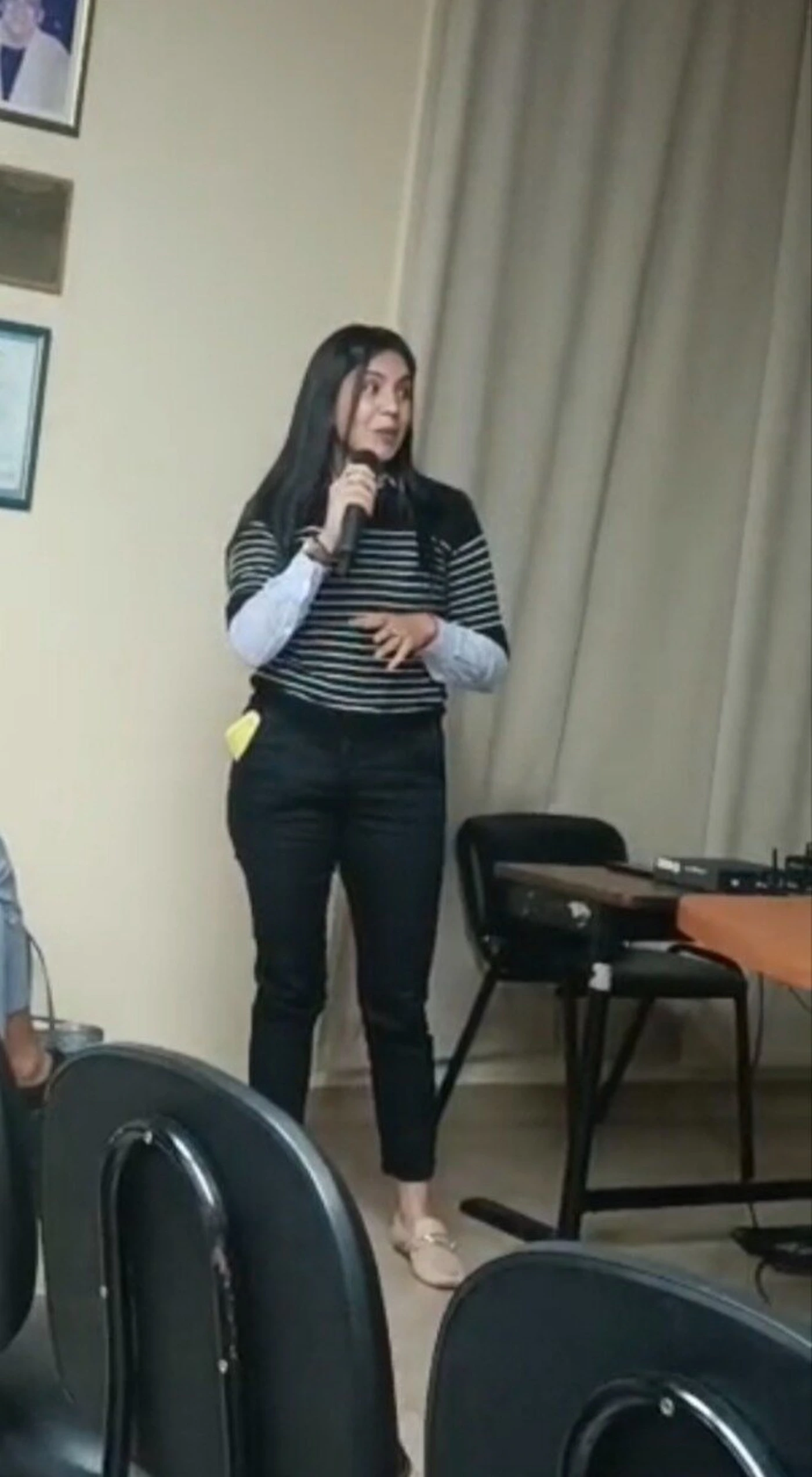 (Photo) A full-shot photograph shows a woman with long dark hair standing indoors, holding a microphone as if speaking or presenting. She is wearing a striped top over a light blue collared shirt, black pants, and light-colored flat shoes, with chairs in the foreground and a table with equipment on the right. person, indoor event | indoor, possibly a presentation or meeting room | woman holding a microphone, framed pictures on the wall (unclear content), chairs in the foreground, table with equipment | candid, documentary | Colors: #E0DCCF, #2E2E2E, #BDBDBD, #D4C5B3 Note: The image captures a real-life scene featuring a person and objects in an indoor setting, which is characteristic of a photograph.
