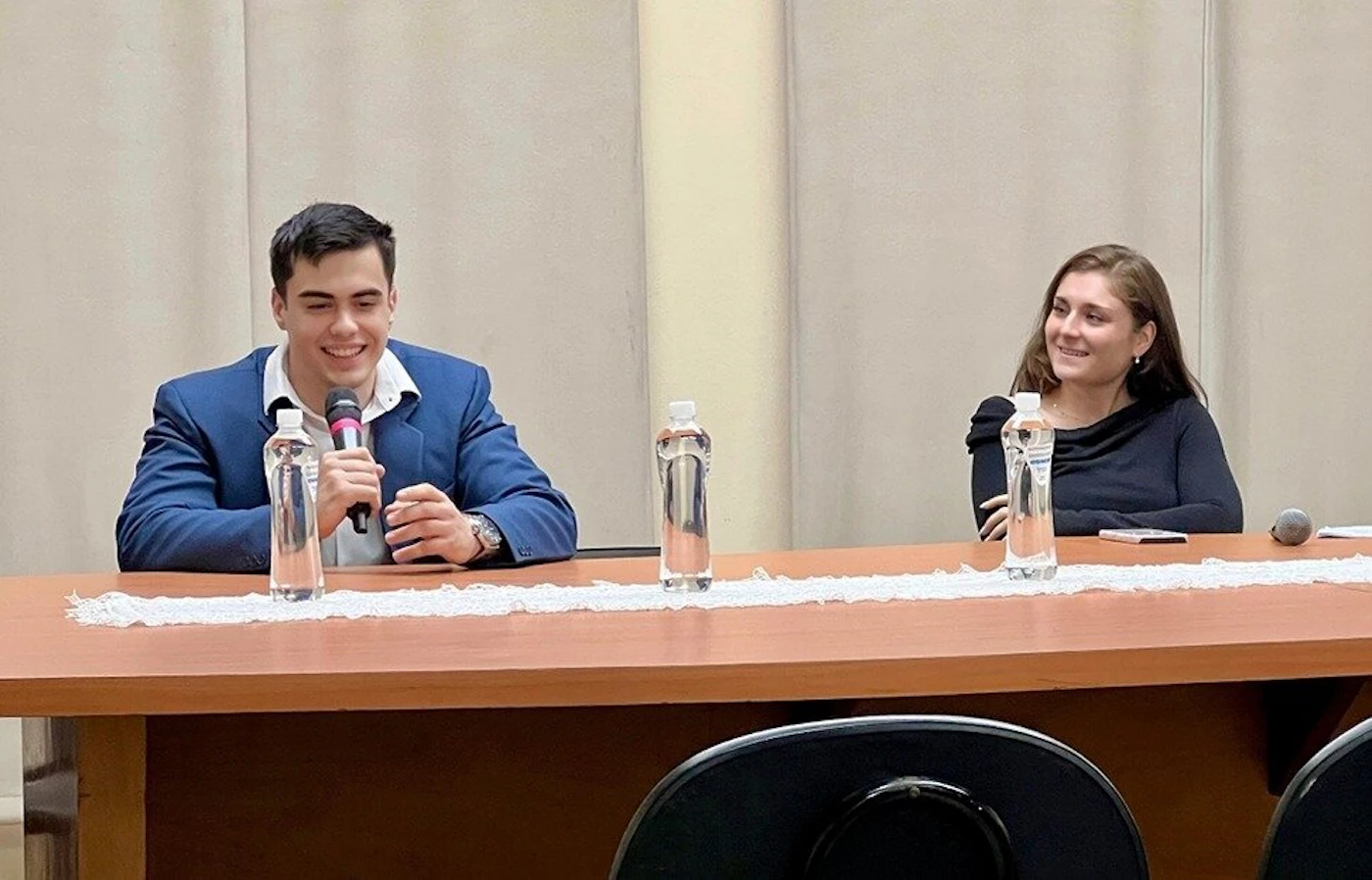 (Photo) The image shows two young adults, a man and a woman, seated at a wooden table with microphones and water bottles, appearing to be in a discussion or presentation setting. The man on the left is holding a microphone and smiling, while the woman on the right is also smiling and looking towards him. people | indoor | two people, microphones, water bottles, wooden table, event/discussion setting | candid | Colors: #3B609E, #2E2C2D, #8B5B3E, #C5C0B7, #FFFFFF Note: This image is a realistic capture of a scene featuring people and objects in a specific setting, which perfectly fits the definition of a 'photo'. It is not a drawing, logo, banner, or handwriting.