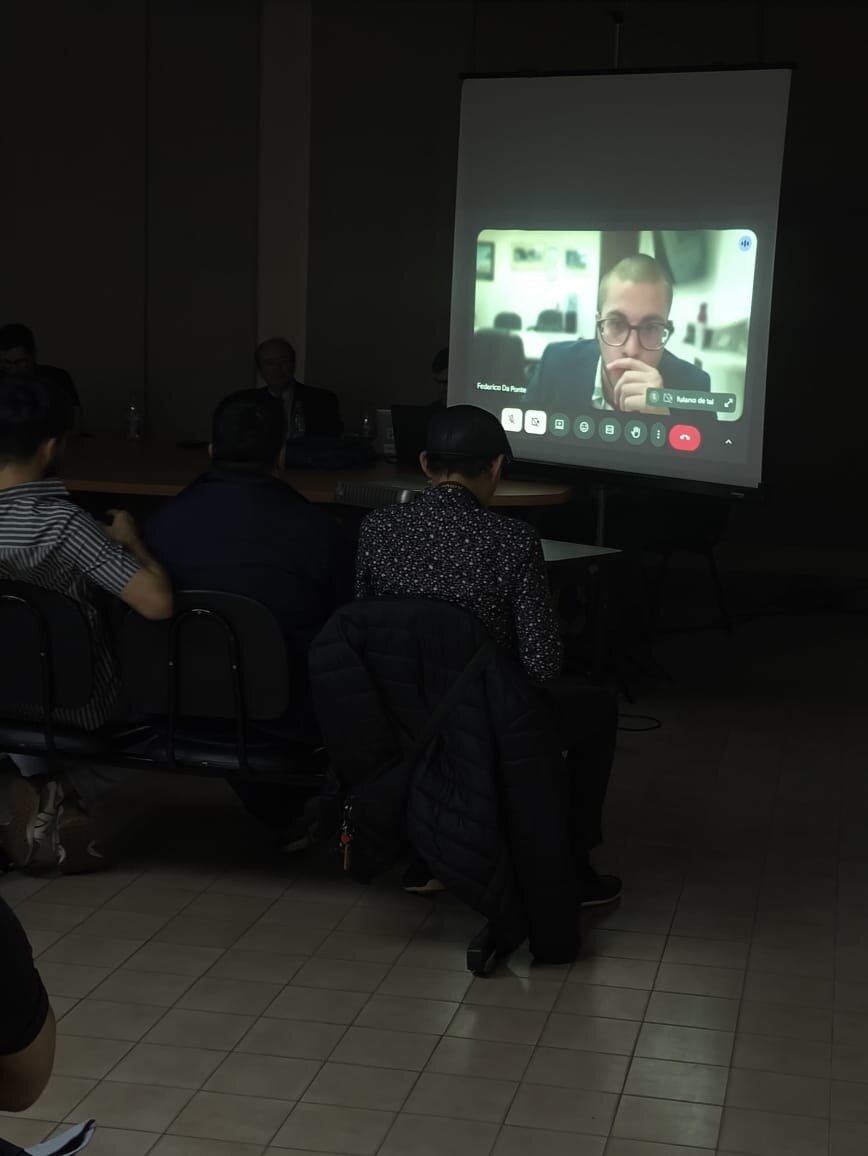 (Photo) The image captures a dimly lit room where several individuals are seated, facing a large projection screen displaying a video conference with a man wearing glasses. The room appears to be a lecture hall or conference room during a presentation. Text: Federico De Ponte, Alumni de [unreadable] People attending a presentation/video conference | Indoor, a dark conference or lecture room | A large projection screen showing a video call, several people seated with their backs to the camera, dim lighting, tiled floor. | Candid, documentary-like | Colors: #2F2F2F, #8D8D8D, #E0F2F1, #3F51B5, #F0C4B4 Note: The image is a realistic capture of a real-world scene, depicting people and a projected screen in a room, characteristic of a photograph rather than an illustration, logo, or banner.