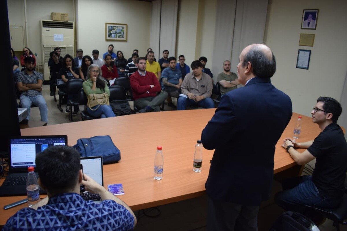 (Photo) The image depicts an indoor scene where a group of people are seated in a room, appearing to attend a lecture or presentation. A man in a dark suit stands at the front, facing away from the camera, addressing the seated audience, while laptops and water bottles are visible on a large wooden table in the foreground. people attending a presentation/lecture | indoor, conference room or classroom | multiple people, a speaker, laptops, water bottles, framed pictures on the wall, air conditioning unit | candid, documentary | Colors: #e0dcd0, #bc724d, #2e3a5a, #8b9a73, #8b2b2b Note: This image is categorized as a 'photo' because it is a direct capture of a real-world event featuring people and objects in a specific setting, rather than an illustration, logo, banner, or handwriting.