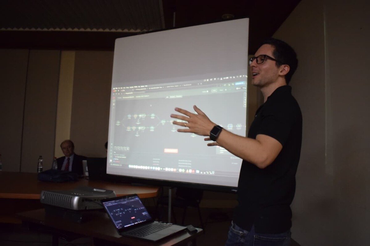 (Photo) A man wearing glasses and a black polo shirt is giving a presentation, gesturing with his hands, in front of a large projection screen displaying a network diagram or technical interface. In the background, another person is partially visible seated at a table, and a projector along with a laptop are on a table in the foreground. Text: [unreadable] A presenter giving a technical presentation or lecture. | Indoor, likely a conference room, lecture hall, or classroom. | A presenter actively gesturing, a large projection screen displaying a network diagram, a partially visible audience member, a projector, and a laptop. | Candid, documentary style, capturing an event. | Colors: #5C5751, #F0F0F0, #1A1A1A, #2B2B2B, #8A5E3F Note: The image is a direct capture of a real-world scene, featuring people, equipment, and an indoor environment, consistent with the definition of a photograph.