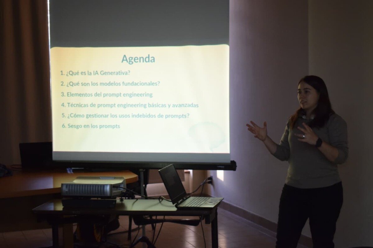 (Photo) The image captures a presentation in progress, featuring a woman gesturing while speaking in front of a large projector screen that displays an agenda in Spanish. Laptops and a projector are visible on a table in the foreground. Text: Agenda 1. ¿Qué es la IA Generativa? 2. ¿Qué son los modelos fundacionales? 3. Elementos del prompt engineering 4. Técnicas de prompt engineering básicas y avanzadas 5. ¿Cómo gestionar los usos indebidos de prompts? 6. Sesgo en los prompts A person giving a presentation | Indoor, likely a conference room or classroom | A female presenter, a projector screen displaying a presentation slide, a projector, and laptops on a table. | Candid, documentary | Colors: #F8F8F8, #8C8C8C, #1A1A1A, #4A2E1D Note: This image is a direct capture of a real-world scene, depicting a person giving a presentation, which classifies it as a photograph rather than an illustration, logo, banner, or handwriting.