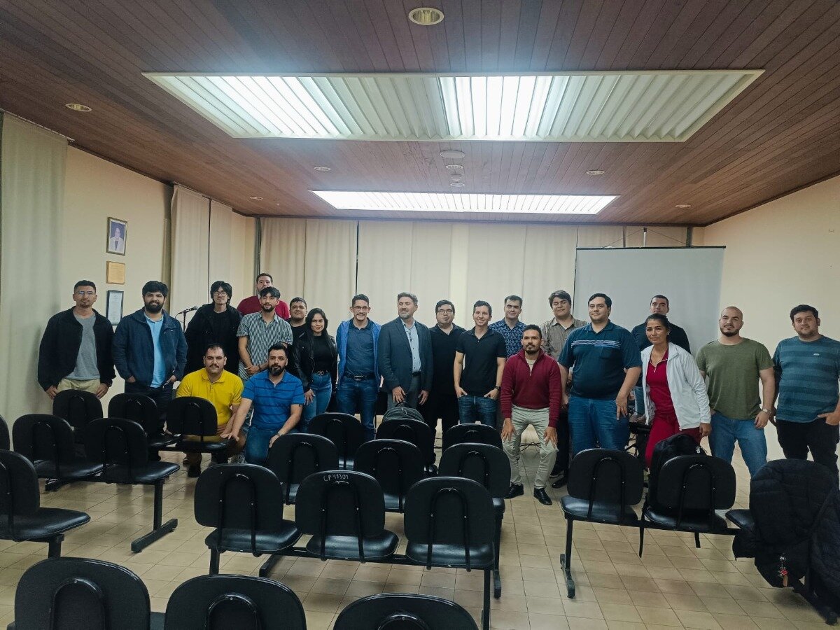 A group of approximately two dozen people, predominantly men, are posing for a photograph in a meeting room or classroom setting. Rows of empty black chairs are visible in the foreground, and the room features a wooden paneled ceiling with light fixtures and a white projection screen on the back wall.. Text: CP 13321