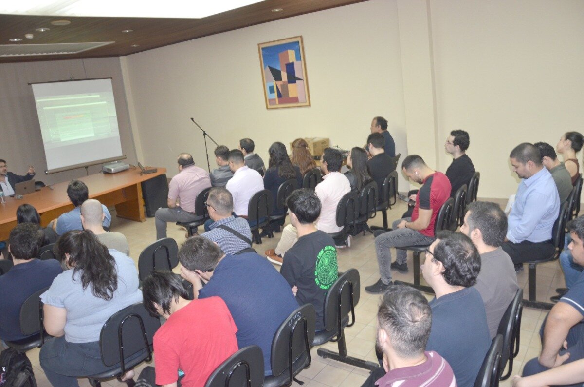 A photograph showing a group of people seated in a lecture hall or conference room, attending a presentation where a speaker is visible on the left next to a projection screen.