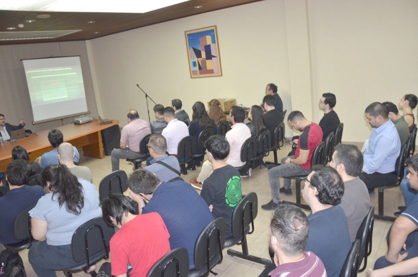 (Photo) A photograph showing a group of people seated in a lecture hall or conference room, attending a presentation where a speaker is visible on the left next to a projection screen. A group of people attending a lecture or presentation. | Indoor, likely a conference room or lecture hall with light walls and wood accents. | An audience is seated facing a presenter; a projection screen is displaying data; abstract artwork hangs on the wall. | Documentary or candid style, taken from the side/back of the audience. | Colors: #EAE3D3, #1A1A1A, #8B4513, #D3D3D3 Note: The image is a realistic capture of a real-world scene, specifically documenting an event or meeting taking place indoors.